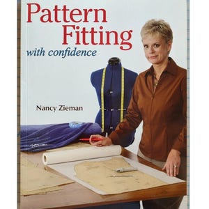 May include: A book titled "Pattern Fitting with confidence" by Nancy Zieman. The cover features the title in red, a woman in a brown shirt, and a blue dress form with a yellow measuring tape. Sewing patterns are on a table.