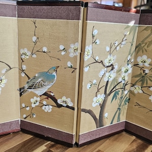 May include: A four-panel folding screen with an Asian-inspired design. Each panel displays a beige backdrop with a painted scene of white blossoms, green bamboo, and a blue and grey bird on a branch. The frame is dark brown.