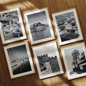May include: A set of six rectangular wall art prints in light wood frames, each featuring a distinct black and white photograph. The images depict various coastal scenes, including a town, a harbor with sailboats, a stone fortress, and palm trees.