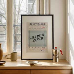 May include: A framed print of a vintage-style guest check with the text "MEET ME IN CARSON" in a black frame. The print is displayed on a wooden surface next to a coffee maker and a mug.