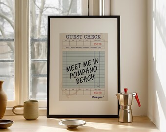 Peut inclure: Une impression encadrée d'un reçu de restaurant de style vintage avec le texte "MEET ME IN POMPANO BEACH". L'impression est dans un cadre noir et repose sur une surface en bois. Une cafetière et une tasse sont également visibles.