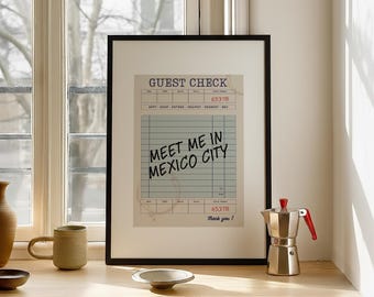May include: A framed print of a guest check with the text "MEET ME IN MEXICO CITY". The print is in a black frame and is displayed on a wooden surface. A coffee maker and a mug are also visible.