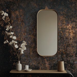 May include: A modern bathroom or entryway scene featuring a gold-framed, oval mirror. Below, a floating shelf displays a white vase with flowers, a candle, and a wooden vase. The wall has a textured, dark brown and gold finish.