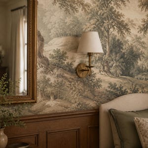 May include: A brass wall sconce with a conical off-white fabric shade mounted on a wall covered in sepia-toned landscape wallpaper. The fixture features a circular backplate, a slender arm, and a small candle-style drip tray beneath the bulb.