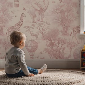 May include: A child sits on a round rug, gazing at a pink and white safari-themed wallpaper. The wallpaper features giraffes, elephants, hot air balloons, and trees. The scene evokes a sense of adventure and imagination.