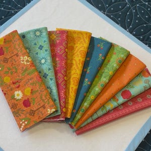 Könnte beinhalten: Fächerförmig angeordnete Stoffmuster mit floralen und Schlüsselmustern in Orange, Türkis, Pink, Gelb und Grün. Die Stoffe liegen auf einer weißen Oberfläche mit blauem Rand.