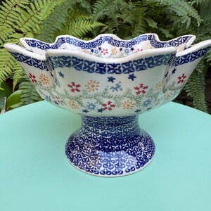 Op de afbeelding: Een witte keramische kom met een blauw en wit bloemenpatroon. De kom heeft een gegolfde rand en staat op een voet.