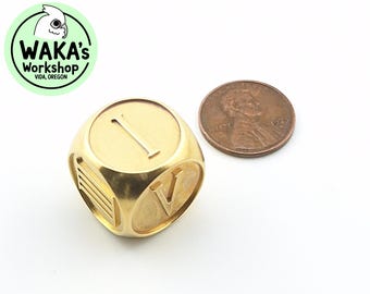 Könnte beinhalten: Ein goldfarbener Würfel aus Metall mit der Zahl eins und der römischen Ziffer V. Der Würfel liegt neben einem US-Penny zum Größenvergleich.