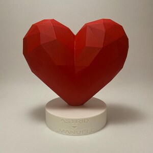 Può includere: Una scultura a forma di cuore geometrico rosso su una base cilindrica bianca. La base presenta i nomi "ASTRID" e "EMANUELE" incisi. Il cuore è di un rosso vibrante.