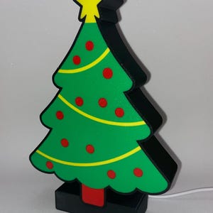 Può includere: Una luce a forma di albero di Natale verde con ornamenti a pois rossi, ghirlanda gialla e una stella gialla in cima. L'albero ha un contorno nero e poggia su una base nera con un accento rosso. Un cavo bianco si estende dalla base.