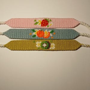 Peut inclure: Trois bracelets d'amitié avec des motifs de fruits différents. Le bracelet du haut est rose avec un motif de fraise, le bracelet du milieu est bleu avec un motif d'orange et le bracelet du bas est vert avec un motif de kiwi.