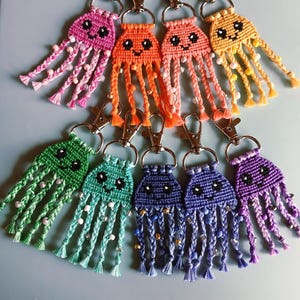 May include: A collection of handmade macrame jellyfish keychains in various colors, including pink, orange, yellow, green, blue, and purple. Each keychain features a smiling face and braided tentacles adorned with beads, attached to a silver-tone clasp.