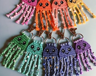 Può includere: Una collezione di portachiavi a forma di medusa fatti a mano in macramè in vari colori, tra cui rosa, arancione, giallo, verde, blu e viola. Ogni portachiavi presenta un volto sorridente e tentacoli intrecciati adornati con perline, attaccati a un fermaglio color argento.