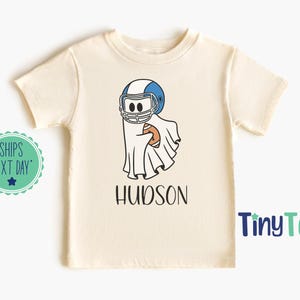 May include: Cream-colored t-shirt featuring a cartoon ghost wearing a blue and white football helmet and holding a football. The name "HUDSON" is printed in black below the graphic. The shirt has short sleeves and a round neckline.