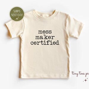 May include: A cream colored short-sleeved t-shirt with the text "mess maker certified" printed in black.