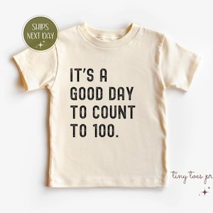 Può includere: T-shirt color crema con la scritta "IT'S A GOOD DAY TO COUNT TO 100" in nero. La maglietta ha maniche corte e girocollo. Una piccola grafica circolare verde e bianca si trova nell'angolo in alto a sinistra.