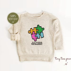 Puede incluir: Una sudadera beige con un diseño de pieza de rompecabezas de colores y el nombre "JAMES" impreso debajo.