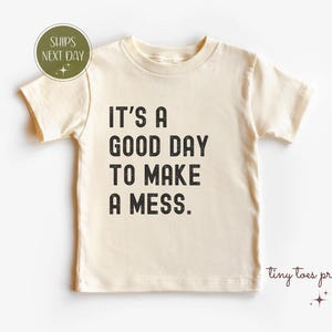 Op de afbeelding: Crèmekleurig peuter-T-shirt met de tekst "IT'S A GOOD DAY TO MAKE A MESS" in het zwart. Het shirt heeft korte mouwen en een ronde halslijn. Een kleine groene cirkel met de tekst "SHIPS NEXT DAY" staat in de linkerbovenhoek.