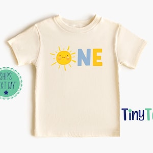 Puede incluir: Camiseta crema de manga corta con un gráfico de sol amarillo sonriente y la palabra "ONE" en azul y amarillo. La camiseta es de TinyToes.