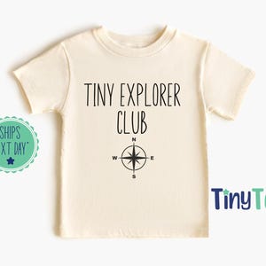 May include: A cream-coloured short-sleeve t-shirt featuring the black text TINY EXPLORER CLUB printed on the front, positioned above a black compass graphic with directional letters N, S, E, and W clearly visible on the design.