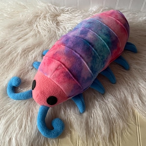 May include: A plush toy shaped like an isopod with a tie-dye pattern in shades of pink, purple, and blue. The creature features a solid pink head, two black circular eyes, and blue antennae and legs extending from its segmented body.
