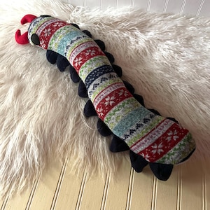 May include: A long, segmented plush toy shaped like a centipede, featuring a colorful knit fabric with red, green, blue, and white snowflake and geometric patterns. It has two bright red antennae, black circular eyes, and many black felt legs.