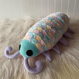 May include: A plush toy shaped like an isopod with a teal head, black circular eyes, and two curled antennae. The body features a textured, multi-colored pattern of pastel pink, blue, and yellow circles, with several lavender-colored legs.