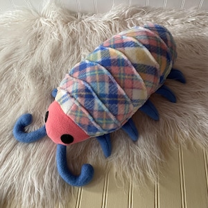 May include: A plush toy shaped like a pill bug with a pink head, black circular eyes, and blue antennae. The segmented body features a multi-colored plaid pattern in shades of blue, pink, yellow, and white, with several blue legs extending outward.