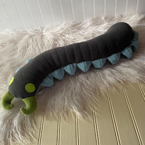 May include: A long, segmented plush toy shaped like a centipede. The body is dark charcoal gray with light blue triangular legs along the sides. It features two bright lime green pincers at the front and two circular lime green eyes.