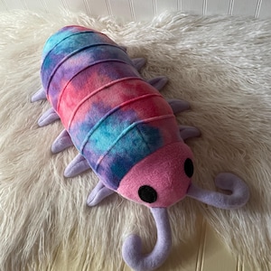 May include: A plush toy shaped like an isopod with a tie-dye pattern in shades of pink, purple, and blue. The segmented body features multiple light purple legs, a solid pink head, two round black eyes, and two long, curved light purple antennae.