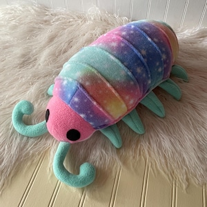 May include: A plush toy shaped like an isopod with a pink head, two black circular eyes, and two curved light blue antennae. The segmented body features a colorful galaxy-inspired print with shades of purple, blue, and pink with white stars.
