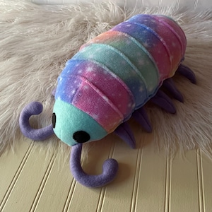 May include: A plush toy shaped like an isopod with a segmented body featuring a rainbow gradient pattern with white star accents. The head is light blue with two large black circular eyes and two purple antennae, and it has multiple purple legs.