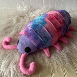 May include: A plush toy shaped like an isopod with a soft lavender head and two black circular eyes. The segmented body features a tie-dye pattern in shades of blue, purple, and pink. It has several pink legs and two curved pink antennae.