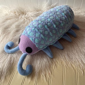 May include: A plush toy in the shape of a bug, featuring a segmented body in shades of blue and turquoise with a textured pattern. The head is lavender with black eyes and blue antennae. The toy has blue legs and is resting on a white, fluffy surface.