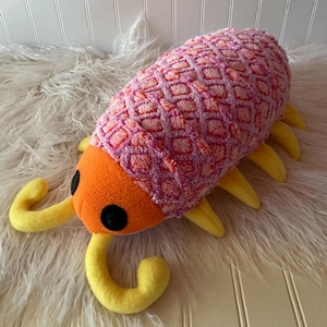 May include: A plush toy shaped like an isopod with a bright orange head, two black circular eyes, and yellow antennae and legs. The body features a textured, diamond-patterned fabric in shades of pink and light purple, creating a soft appearance.