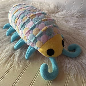 May include: A plush toy shaped like an isopod with a segmented body covered in a soft, fuzzy pattern of pastel pink, blue, and yellow circles. It features a solid yellow head, two large black circular eyes, and light blue curled antennae and legs.
