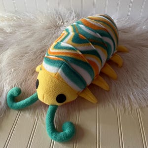 May include: A plush isopod toy featuring a yellow head with black circular eyes and two teal antennae. The segmented body is covered in a swirling pattern of orange, teal, and white stripes, with multiple yellow legs extending from the sides.