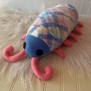 May include: A plush toy shaped like an isopod with a blue head, two black circular eyes, and two curved pink antennae. The body features a soft, multi-colored plaid pattern in shades of blue, pink, and yellow, with several pink legs.