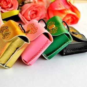 Puede incluir: Cuatro pequeñas bolsas de cuero en oro, rosa, verde y negro. Cada bolsa tiene una solapa con un diseño dorado y un pequeño cordón negro. Las bolsas están dispuestas en fila, con un fondo de rosas rosas y rojas.