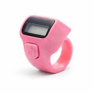 May include: A pink digital finger counter with a black display screen and a button with a curved design. The counter is ring-shaped and made of plastic. The device is designed to be worn on a finger.