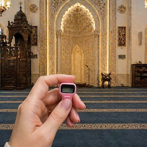 May include: A pink digital counter showing the number 15. The counter is held in front of a mosque interior with ornate architectural details, including a decorated archway and patterned carpet.