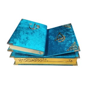 Op de afbeelding: Een stapel van drie boeken met teal fluwelen covers en gouden accenten. Het open boek toont gouden Arabische kalligrafie en een gouden embleem. De pagina randen zijn goud, en de hoeken zijn versterkt met goud metaal.