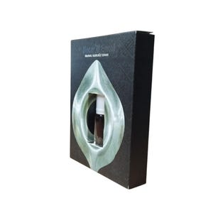 May include: A black box with a silver design and the text "Hajar Al-Aswad" and "Original Al-Khususi E-Dans". A small glass bottle of perfume is visible through a cutout in the box.