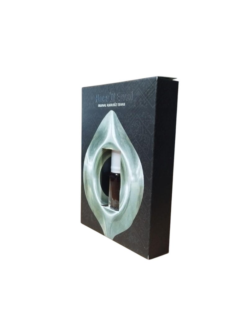 May include: A black box with a silver design and the text "Hajar Al-Aswad" and "Original Al-Khususi E-Dans". A small glass bottle of perfume is visible through a cutout in the box.