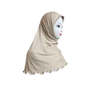May include: A beige hijab, a head covering worn by some Muslim women. The hijab is draped over a mannequin head, showcasing its design. The fabric has a smooth texture and a scalloped edge with white trim.