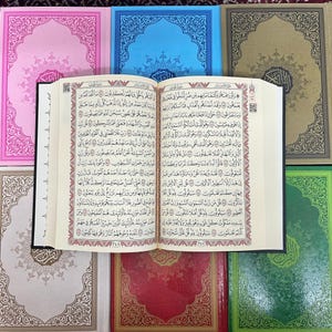 May include: An open Quran with Arabic text in black and red ink on white paper. The pages are bound in black and the cover is white with a decorative border. The book is surrounded by six smaller books with decorative covers in various colors, including pink, blue, green, and red.