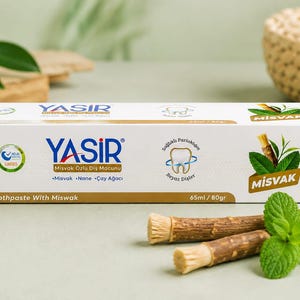 Könnte beinhalten: Eine rechteckige weiße Schachtel Yasir Zahnpasta mit Miswak, Minze und Teebaumöl. Die Verpackung zeigt den Markennamen, eine Zahngrafik und den Text Toothpaste With Miswak, 65ml / 80gr. Zwei Miswak-Stäbchen und frische Minzblätter liegen vor der Schachtel.