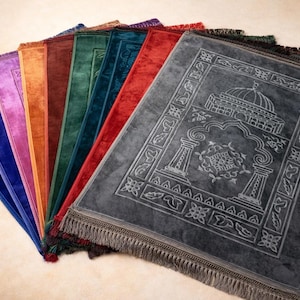Op de afbeelding: Een verzameling gebedskleden in verschillende kleuren, waaronder blauw, paars, oranje, bruin, groen, blauwgroen en rood. De kleden zijn rechthoekig met decoratieve randen en franjes. Het voorste kleed is grijs met een gedetailleerd ontwerp.