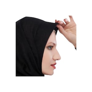 Può includere: Un hijab nero, un copricapo tradizionale, è mostrato di profilo. Una mano regola il bordo dell'hijab. Il viso della donna è visibile, con pelle chiara e trucco scuro per gli occhi.