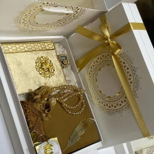 May include: A white gift box with gold detailing, containing a gold-coloured book, a beaded necklace, and a decorative feather. The box is tied with a gold ribbon. The lid of the box features a decorative lace pattern.